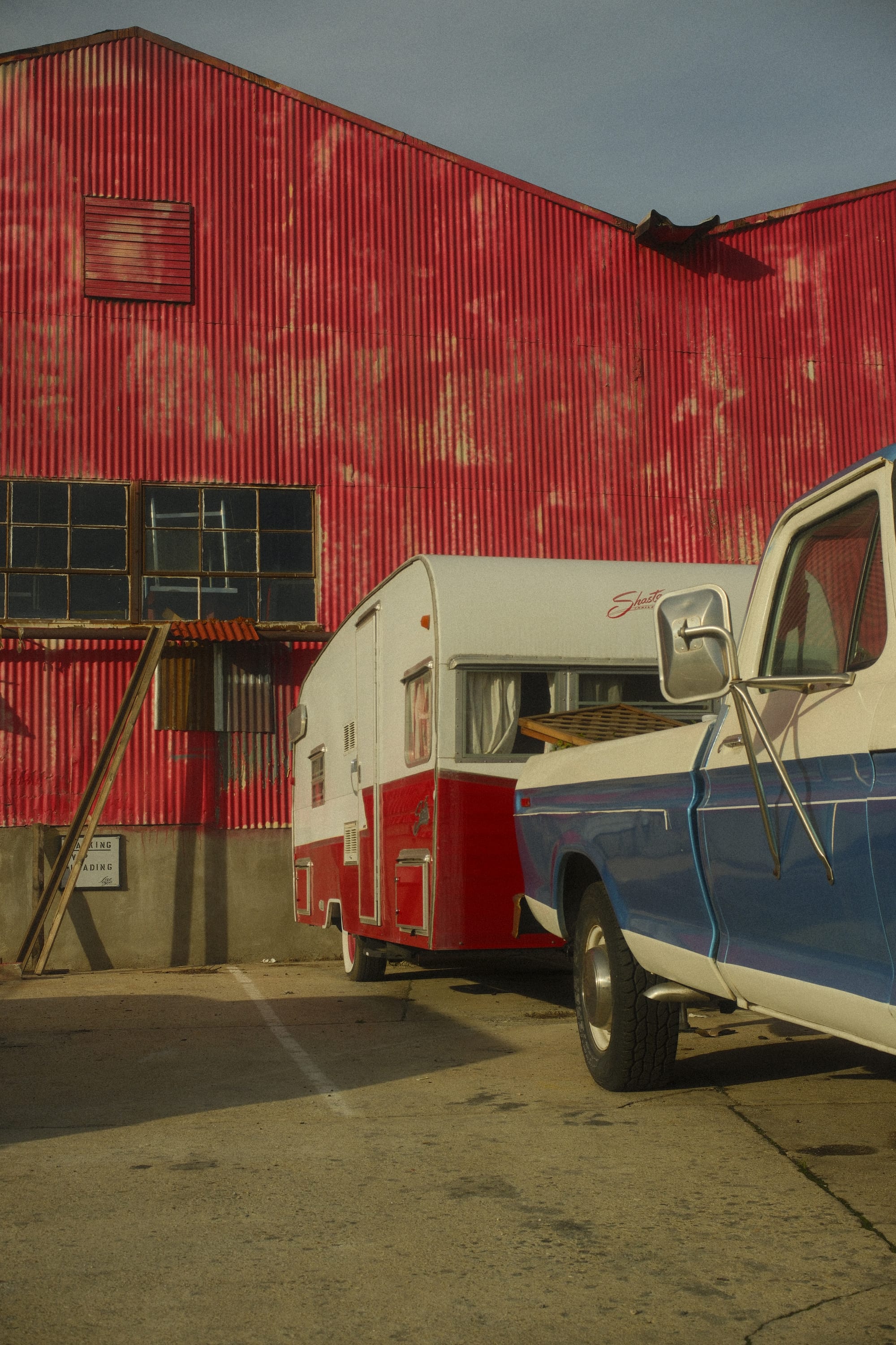 a blue and white truck has a red and white trailer attached to it. these are both in front of a big red corrugated steel building.